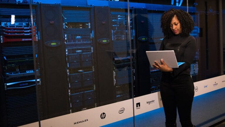 Mujer trabajando con un portátil frente a servidores en un centro de datos. Representa soluciones tecnológicas ofrecidas por compañías de software.