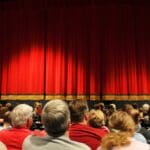 Grupo de personas disfrutando de una función de teatro, con el auditorio iluminado y la atención centrada en el escenario.