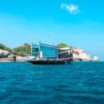 Barco turístico navegando en aguas cristalinas cerca de una isla rocosa con exuberante vegetación bajo un cielo azul despejado.