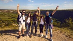 Group of five young hikers with backpacks smiling and raising their arms on top of a hill, overlooking a scenic valley with green fields and mountains under a clear blue sky.