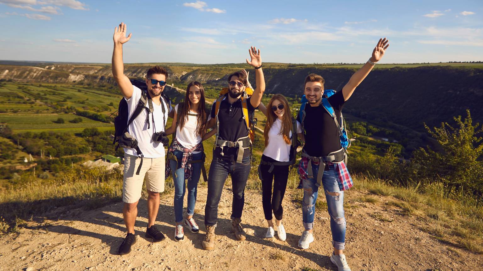 Group of five young hikers with backpacks smiling and raising their arms on top of a hill, overlooking a scenic valley with green fields and mountains under a clear blue sky.