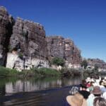 Grupo de turistas navegando por un río rodeado de imponentes acantilados rocosos bajo un cielo azul despejado en un paisaje natural impresionante.