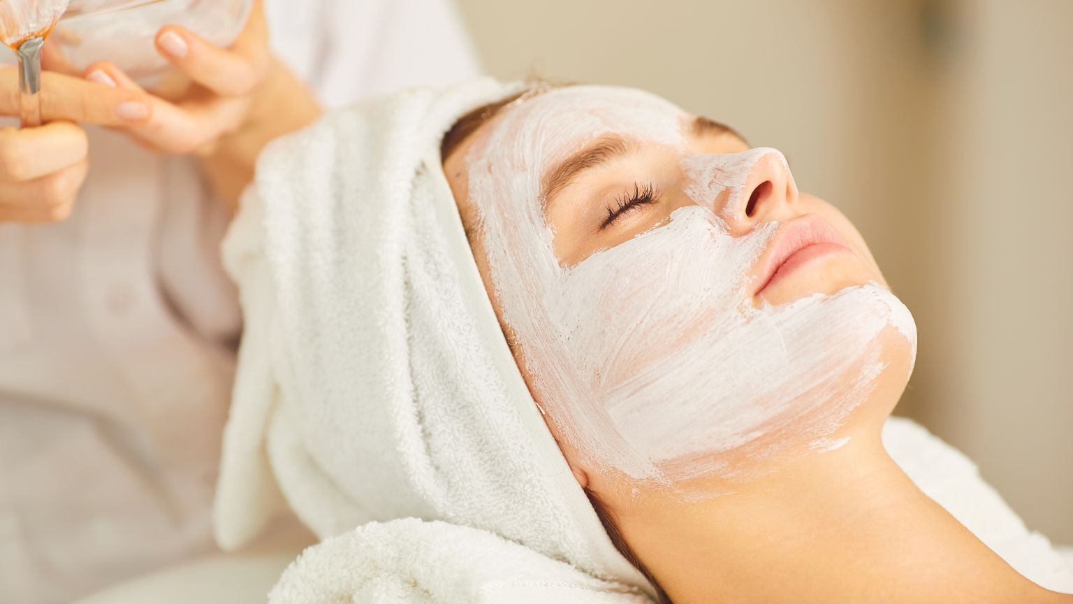 Mujer relajada recibiendo un tratamiento facial en un centro de estética, con toalla en la cabeza y productos hidratantes en su piel.