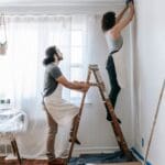 Pareja realizando una reforma en casa, pintando y renovando una habitación con herramientas y escalera.