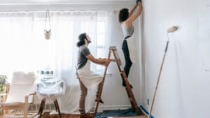 Pareja realizando una reforma en casa, pintando y renovando una habitación con herramientas y escalera.