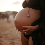 Mujer embarazada en sesión de fotos con luz natural, sonriendo y abrazando su barriga en un entorno emocional y cálido.