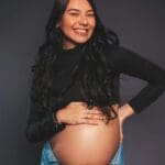 Mujer embarazada en estudio de fotografía profesional con luz suave, posando de perfil y sonriendo mientras sostiene su barriga