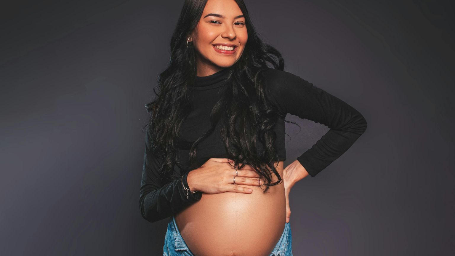 Mujer embarazada en estudio de fotografía profesional con luz suave, posando de perfil y sonriendo mientras sostiene su barriga