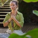 Mujer practicando mindfulness en la naturaleza, sentada en calma con los ojos cerrados, representando meditación guiada, atención plena y reducción del estrés.