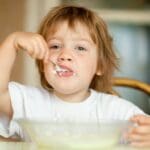 Niño pequeño comiendo yogur con cuchara en la mesa durante el desayuno.