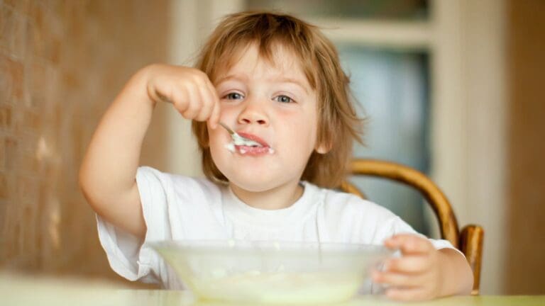 Niño pequeño comiendo yogur con cuchara en la mesa durante el desayuno.