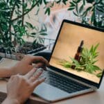 Persona comprando aceite CBD online desde su ordenador en España, junto a productos naturales y certificados sobre una mesa de madera.