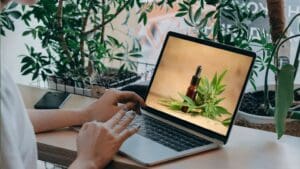 Persona comprando aceite CBD online desde su ordenador en España, junto a productos naturales y certificados sobre una mesa de madera.
