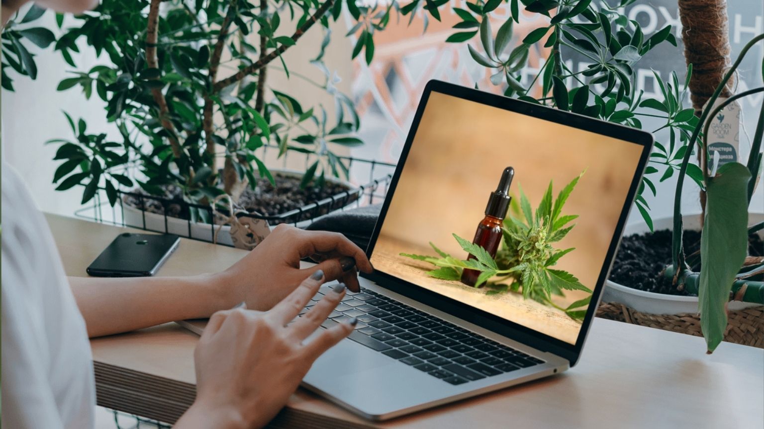 Persona comprando aceite CBD online desde su ordenador en España, junto a productos naturales y certificados sobre una mesa de madera.