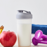 Frutas, batido proteico, mancuernas y toallas sobre una mesa como símbolo de hábitos saludables y entrenamiento en casa.