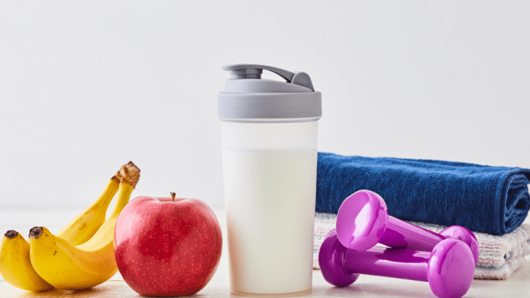 Frutas, batido proteico, mancuernas y toallas sobre una mesa como símbolo de hábitos saludables y entrenamiento en casa.