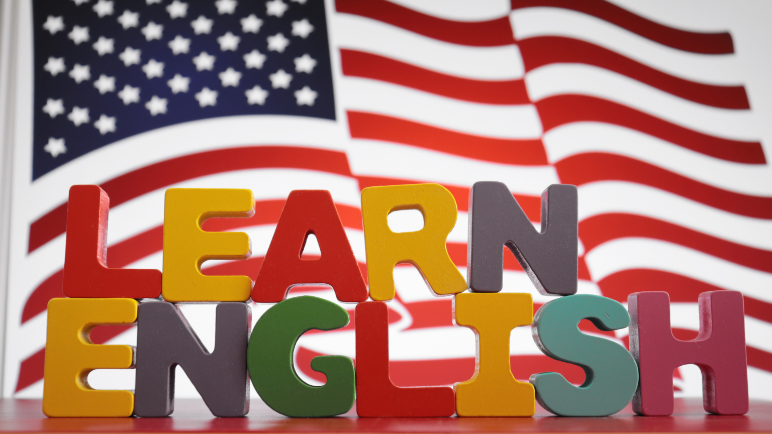 Letras de colores formando la frase "Learn English" frente a una bandera de Estados Unidos como símbolo del aprendizaje del inglés en un contexto lúdico y educativo.