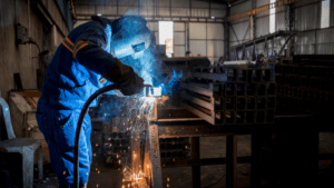 Soldador profesional trabajando en taller industrial con estructura metálica, aplicando técnica de soldadura MIG en tubos de acero.