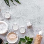 Productos de cosmética natural en envases neutros junto a hojas verdes sobre fondo claro minimalista.