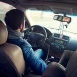 Alumno durante clases prácticas de conducir en ciudad, vista desde el asiento trasero del vehículo con el conductor al volante y circulación urbana al fondo