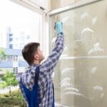 Hombre realizando limpieza profesional de cristales en ventana exterior con herramientas especializadas.