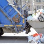 camión de recogida de residuos vaciando contenedor con bolsas de basura acumuladas en zona urbana