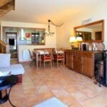 Salón de casa rural con chimenea cerrada, sofá amplio, mesa de comedor de madera y escalera interior en entorno acogedor de montaña.