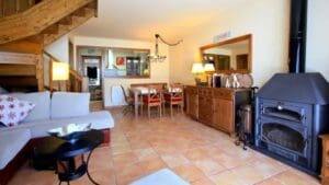 Salón de casa rural con chimenea cerrada, sofá amplio, mesa de comedor de madera y escalera interior en entorno acogedor de montaña.